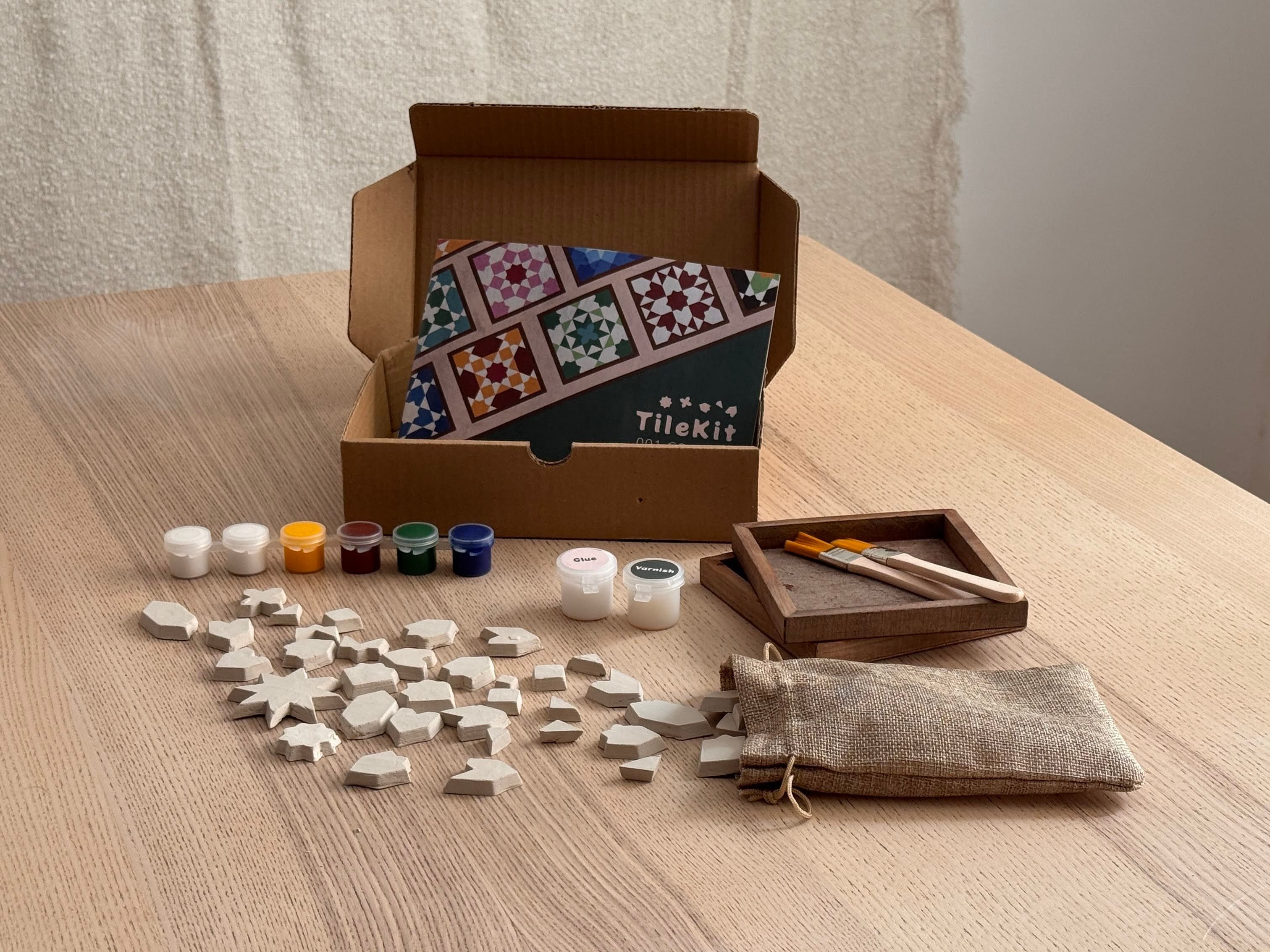 Geöffnete TileKit-Box auf einem Tisch mit Tonfliesen, Farben, Pinseln und Rahmen