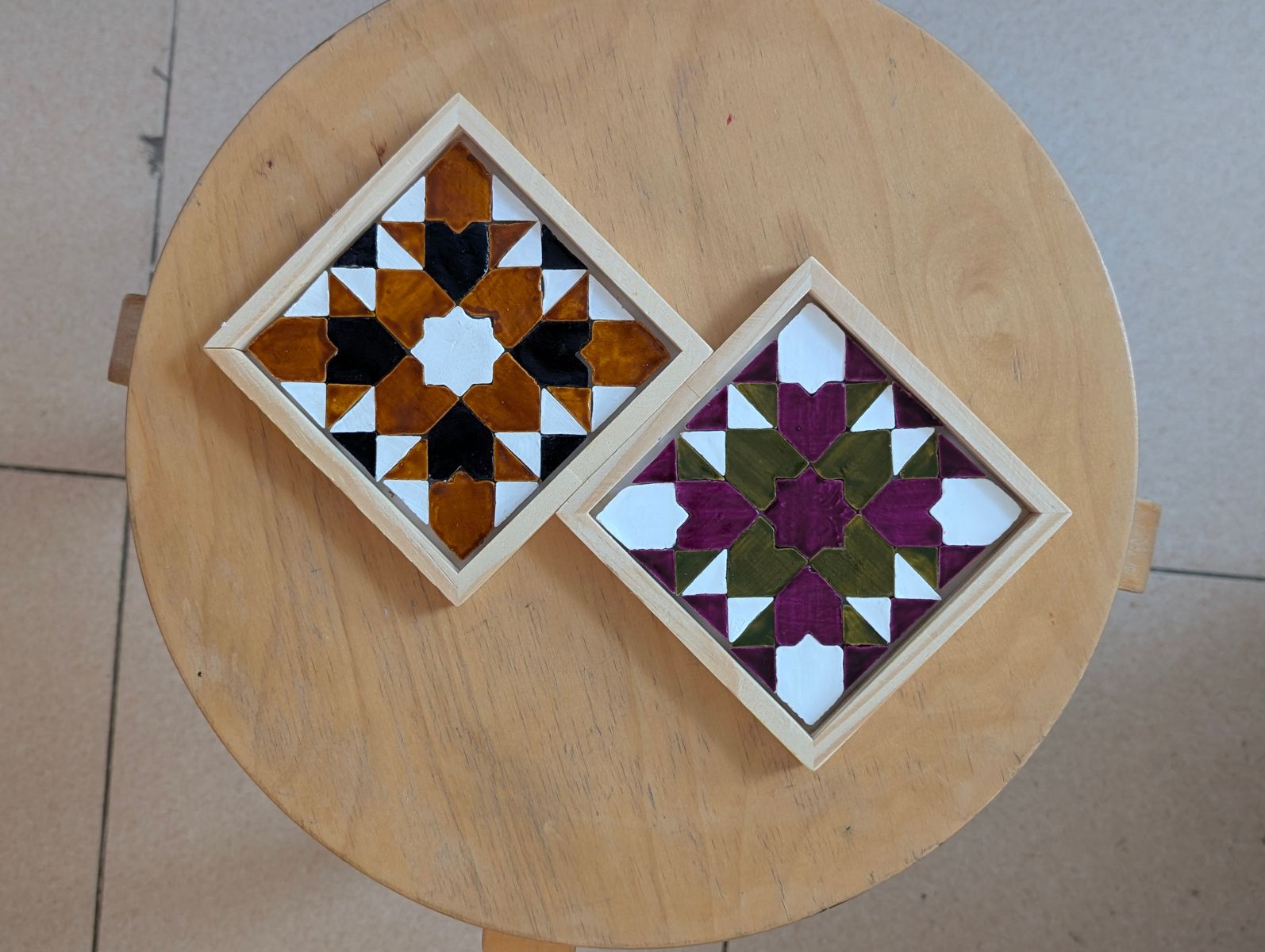Zwei fertige geometrische Mosaik-Untersetzer auf einem runden Holzhocker